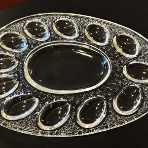 May include: Clear glass serving platter with a central oval section and twelve surrounding oval indentations. The platter has a textured, crackled design around the edges. The platter is on a black surface.