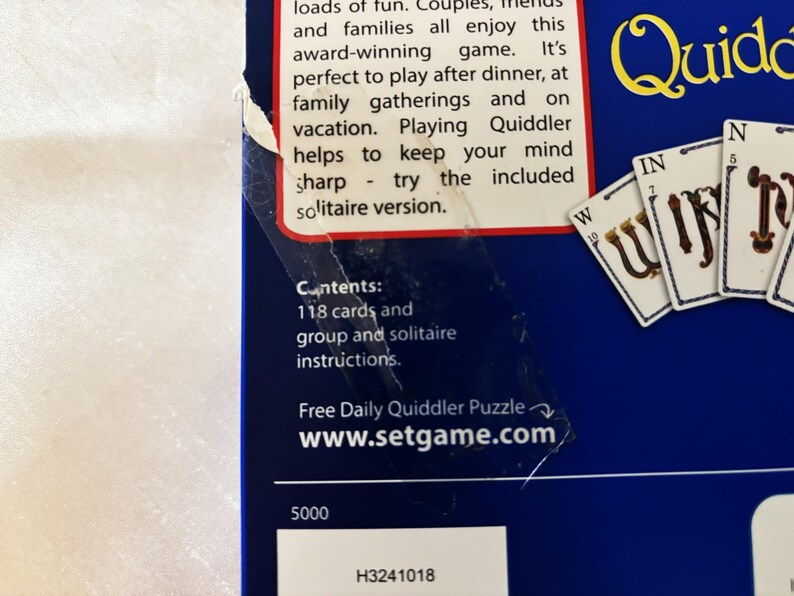 May include: A close-up of a blue game box with the word "Quiddler" in yellow. The box includes text about the game and its contents, including 118 cards and instructions. Several game cards are visible.