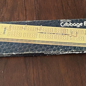 May include: A wooden cribbage board game in its original box. The box is black with white text that reads "Wooden Cribbage Board". The board is made of wood and has a dark brown finish. The board is divided into sections with pegs and scoring areas.