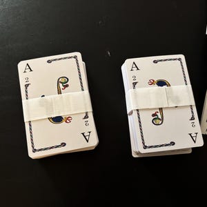 May include: Two decks of playing cards, each secured with a white paper band. The cards feature the Ace of Spades and the number 2, with decorative borders and symbols. The background is a dark surface.