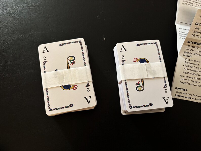 May include: Two decks of playing cards, each secured with a white paper band. The cards feature the Ace of Spades and the number 2, with decorative borders and symbols. The background is a dark surface.