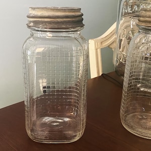 May include: Two clear glass jars with metal lids. The jars have a grid pattern on the glass. The jars are sitting on a wooden surface.