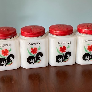 May include: Set of four vintage white glass spice jars with red lids and black and white floral designs. The jars are labeled "Cloves", "Paprika", "Allspice", and "Cinnamon".