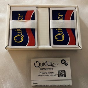 May include: A new Quiddler card game set in a white box. Two stacks of cards are visible, each secured with a white paper band. The card backs are navy blue with a red and white design. A printed instruction sheet is also included.