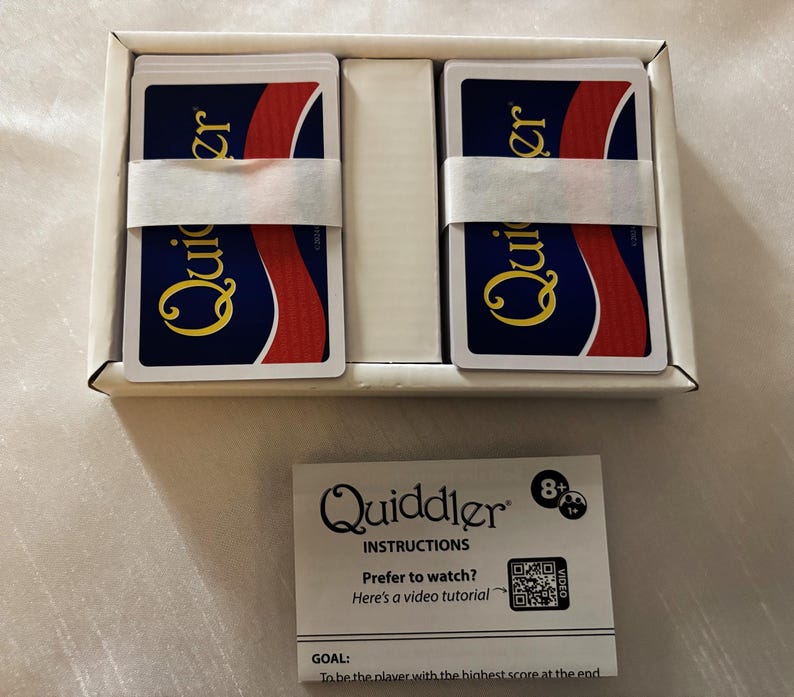 May include: A new Quiddler card game set in a white box. Two stacks of cards are visible, each secured with a white paper band. The card backs are navy blue with a red and white design. A printed instruction sheet is also included.