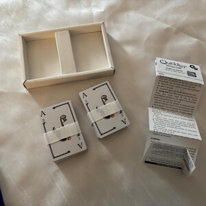 May include: A card game set with two decks of cards, each secured with a white band, and a white box. The cards feature an ace of spades design. Instructions are included. The game is called "Quiddler".