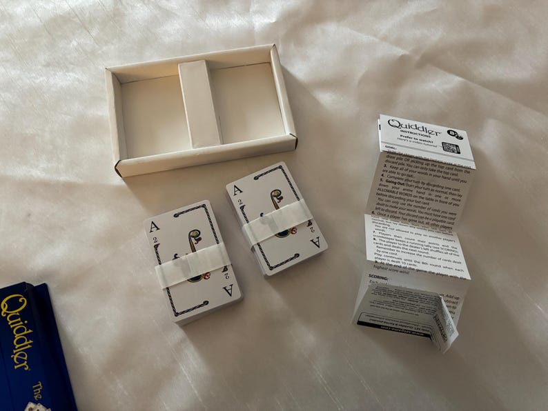 May include: A card game set with two decks of cards, each secured with a white band, and a white box. The cards feature an ace of spades design. Instructions are included. The game is called "Quiddler".