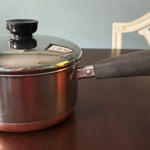 May include: Stainless steel saucepan with a lid and a black handle. The pot has a copper-colored base and a black knob on the lid. A metal ring is attached to the end of the handle. Kitchenware.