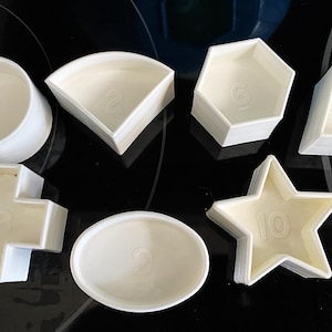 May include: A collection of white plastic cookie cutters in various shapes, including a circle, a quarter circle, a hexagon, a trapezoid, a cross, an oval, and a star. Each cutter has a number embossed on it.
