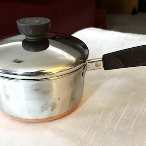 May include: A small, stainless steel saucepan with a lid and a black handle. The pot has a copper-colored bottom and a black knob on the lid. A metal ring is attached to the end of the handle.