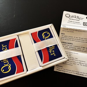 May include: A Quiddler card game set in a white box, with two stacks of cards secured by a white band. The cards have a blue background with a red stripe and the word "Quiddler" in yellow. Instructions are visible.