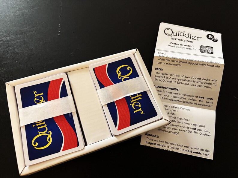 May include: A Quiddler card game set in a white box, with two stacks of cards secured by a white band. The cards have a blue background with a red stripe and the word "Quiddler" in yellow. Instructions are visible.