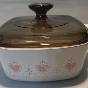 Puede incluir: Cazuela blanca con tapa y asa de cristal marrón. El plato presenta un patrón de corazón y flores rosas. El plato tiene asas blancas a cada lado. Un artículo de cocina vintage.