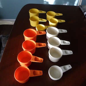 May include: A set of 12 measuring cups in three colors: orange, white, and yellow. The cups are made of plastic and have a handle on one side.