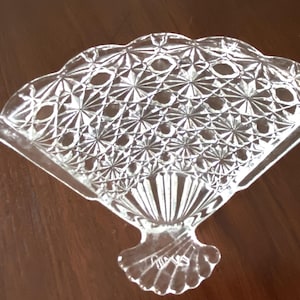 May include: A clear glass fan-shaped napkin holder with a decorative diamond pattern. The holder has a scalloped edge and a small base.