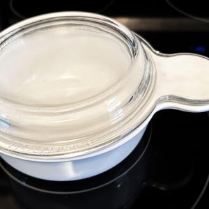 May include: A white and clear glass microwave bowl with a handle. The bowl has a clear glass lid and is sitting on a black stovetop. The bowl is round and has a handle on one side.