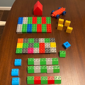 May include: A set of colorful plastic building blocks, including a red house, a yellow car, and various shaped blocks in green, blue, red, yellow, gray, and white.