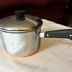May include: A small stainless steel saucepan with a lid and a black handle. The bottom of the pan has a copper band. The handle has a metal loop at the end. The pan is sitting on a white tablecloth.