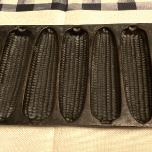 May include: A cast iron corn stick pan with seven corn-shaped molds. The pan is dark gray and has a rectangular shape with handles on each end. The molds are detailed to resemble ears of corn, perfect for baking.