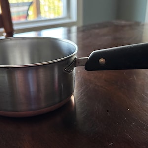 May include: A stainless steel saucepan with a copper-colored base and a black handle. The handle is secured with two metal rivets and features a small metal loop at the end for hanging. The interior of the pan is smooth and metallic silver.