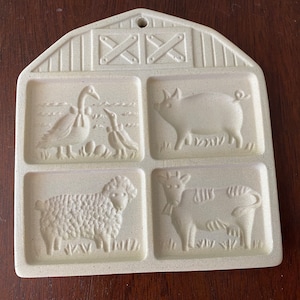 May include: A white ceramic cookie cutter shaped like a barn with four different animal designs: a goose, a pig, a sheep, and a cow.