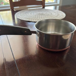 May include: A stainless steel saucepan with a dark gray handle featuring two metal rivets and a circular metal hanging loop at the end. The base of the pan has a copper-colored exterior finish, and the interior is polished silver-toned metal.