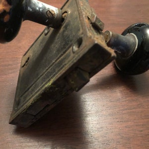 May include: Antique black porcelain door knob set with a metal backplate. The knob is round and has a smooth finish. The backplate is rectangular and has a worn, aged look.