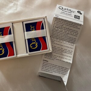 May include: A new Quiddler card game set in a white box. The game includes two stacks of cards, each wrapped with a white band. The cards have a blue background with the word "Quiddler" in gold. Instructions are included.