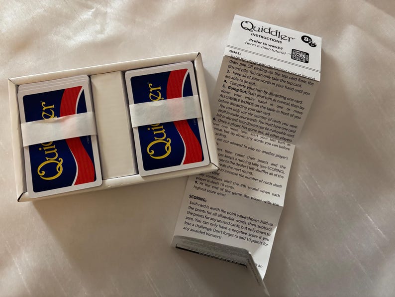 May include: A new Quiddler card game set in a white box. The game includes two stacks of cards, each wrapped with a white band. The cards have a blue background with the word "Quiddler" in gold. Instructions are included.
