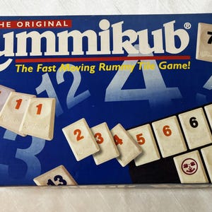 May include: Rummikub game box with the text "THE ORIGINAL Rummikub" and "The Fast Moving Rummy Tile Game!" on a blue background. The box displays game tiles with numbers 1-7 and 13. The game is for 2 to 4 players.