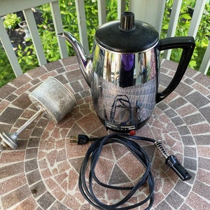 May include: A vintage chrome and black electric coffee percolator with a cord and a separate metal filter. The percolator is sitting on a tiled surface.