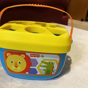 May include: A Fisher-Price shape sorter toy in blue and yellow with an orange handle. The toy features a lion and alligator graphic. The top has multiple shape holes for inserting blocks. The toy is designed for toddlers.