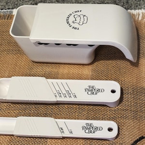 May include: White Pampered Chef kitchen tools, including a measuring cup and two adjustable measuring spoons. The measuring cup has a curved handle and the brand logo. The spoons have measurement markings and the brand name.