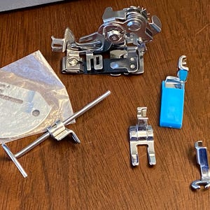 May include: A set of sewing machine feet, including a zipper foot, a blind stitch foot, a walking foot, a buttonhole foot, and a foot for sewing on buttons. The feet are made of metal and have a silver finish. There is also a metal foot attachment bar and a round metal sewing machine foot plate.