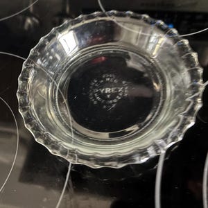 May include: Clear glass pie plate with a fluted edge. The Pyrex brand is visible on the bottom of the plate. The plate is sitting on a black stovetop.