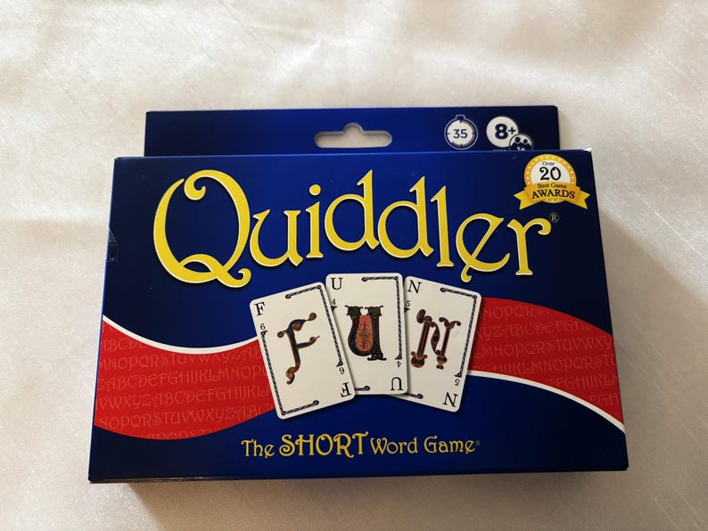 May include: A blue game box with the word "Quiddler" in yellow script. The box features three cards spelling "FUN". The text "The SHORT Word Game" is below the cards. The box also has a "20 Game Awards" seal.