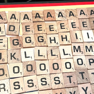 May include: Wooden Scrabble tiles with black letter and number markings. The tiles are arranged on a red and yellow surface, displaying various letters including A, B, D, E, F, G, H, I, J, K, L, M, N, O, P, R, S, T, Y, and Z.