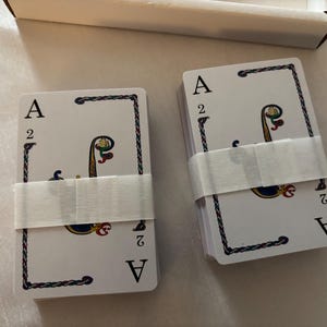 May include: Two decks of playing cards, each wrapped with a white paper band. The cards feature the Ace of Spades and the number 2, with a decorative design in the center. The cards are in a white box.