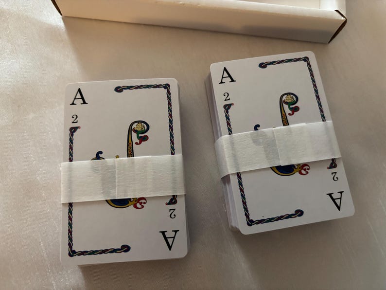 May include: Two decks of playing cards, each wrapped with a white paper band. The cards feature the Ace of Spades and the number 2, with a decorative design in the center. The cards are in a white box.