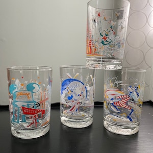 May include: Four clear glass tumblers with colorful illustrations of Disney characters and landmarks. The tumblers feature Mickey Mouse, Donald Duck, Goofy, and the Hollywood Tower Hotel.
