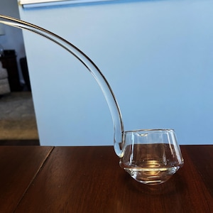 May include: A clear glass ladle with a curved handle and a unique, angular bowl. The ladle is resting on a dark brown wooden surface, with a light blue wall in the background.