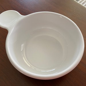 May include: A white ceramic bowl with a rounded shape and a small, circular handle. The bowl is empty and sits on a dark wooden surface. The handle is on the left side of the bowl.