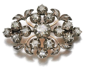 May include: An antique brooch with a floral design. The brooch features numerous clear, round-cut gemstones set in a silver-toned metal frame with gold accents. The gemstones are arranged in a symmetrical pattern, with a large central stone.