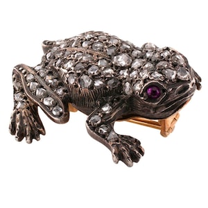 Puede incluir: Un broche de plata en forma de rana con un diseño detallado, cubierto de pequeños diamantes y acentuado con un solo ojo de rubí.