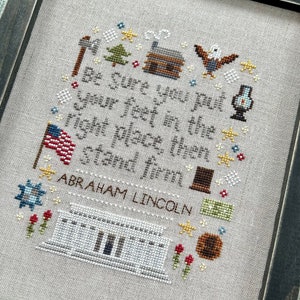 Puede incluir: Obra de arte de punto de cruz en un marco de madera oscura. El diseño presenta una cita de Abraham Lincoln, rodeada de imágenes patrióticas y rústicas, incluyendo una bandera americana, una cabaña de madera y estrellas.