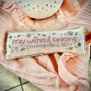 May include: A white pillow with green trim and a cross-stitch design featuring the text "pray without ceasing 1 Thessalonians 5:17" surrounded by colorful flowers.