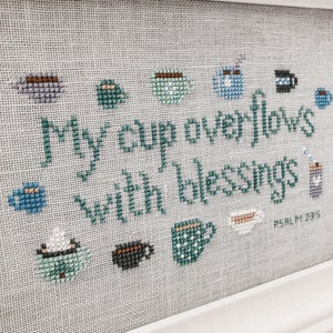 Peut inclure: Broderie au point de croix sur fond blanc avec du fil turquoise et bleu. La broderie représente une variété de tasses à café et le texte "My cup overflows with blessings Psalm 23:5".