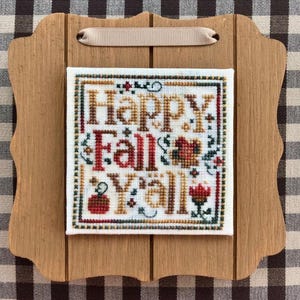 May include: A cross-stitch embroidery on a wooden frame with a tan ribbon. The embroidery says "Happy Fall Y'all" with autumnal colors and designs.