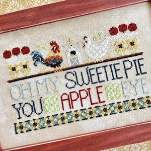 May include: A framed cross-stitch sampler on beige fabric featuring a rooster, a white hen, a small house, red apples, and yellow flowers. The text reads: OH MY SWEETIE PIE YOU APPLE EYE, with a decorative blue and yellow checkered border at the bottom.