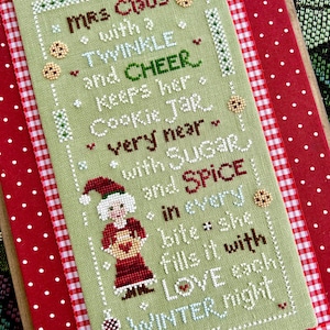 Pode incluir: Obra de arte em ponto cruz com a Sra. Claus e o texto: "Mrs. Claus with a twinkle and cheer keeps her cookie jar very near with sugar and spice in every bite she fills it with love each winter night." A obra de arte está sobre um fundo verde com uma borda de bolinhas vermelhas e brancas.
